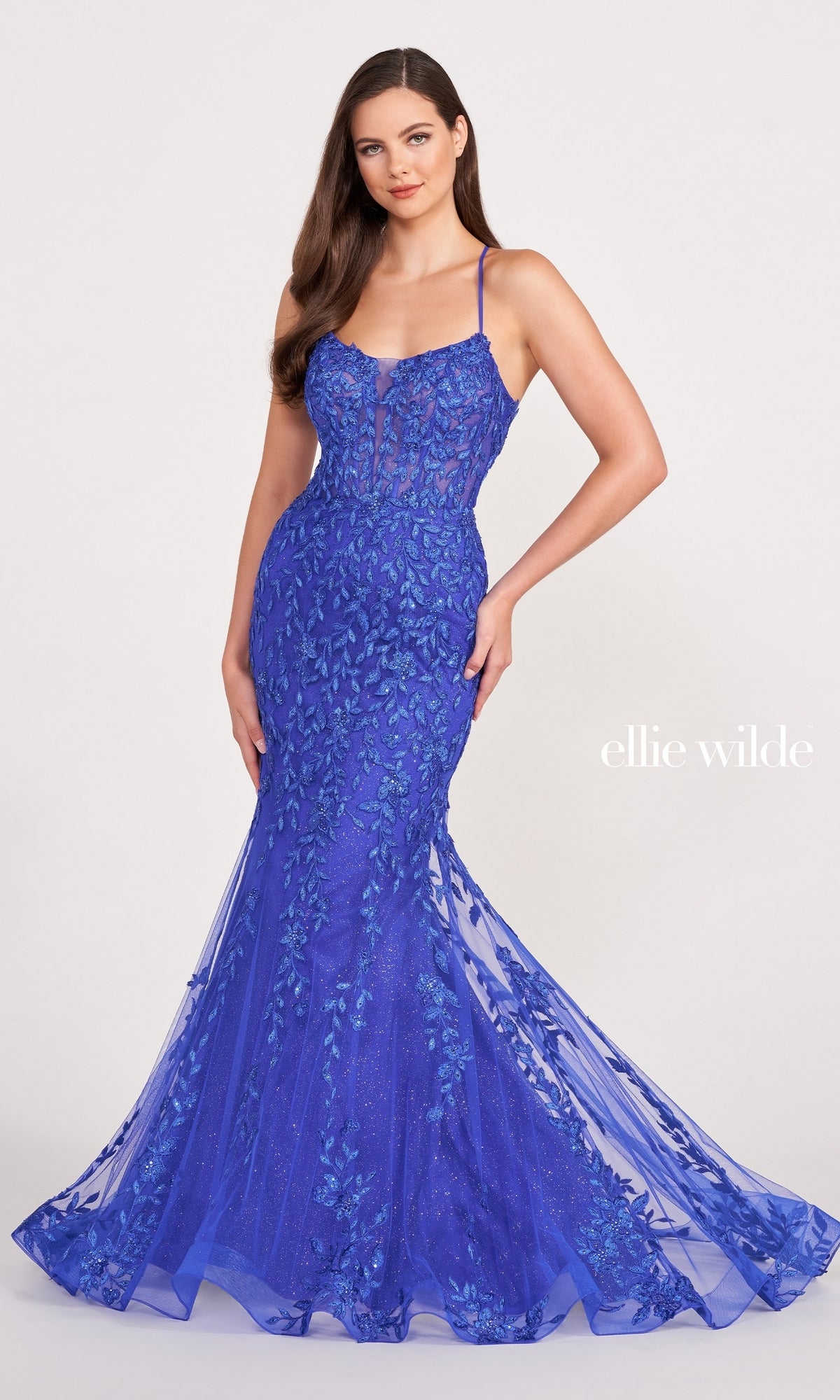 A woman poses against a plain background in the EW34090 Lace Applique Over Glitter Jersey Gown, a fitted, floor-length royal blue mermaid dress with thin straps. The text